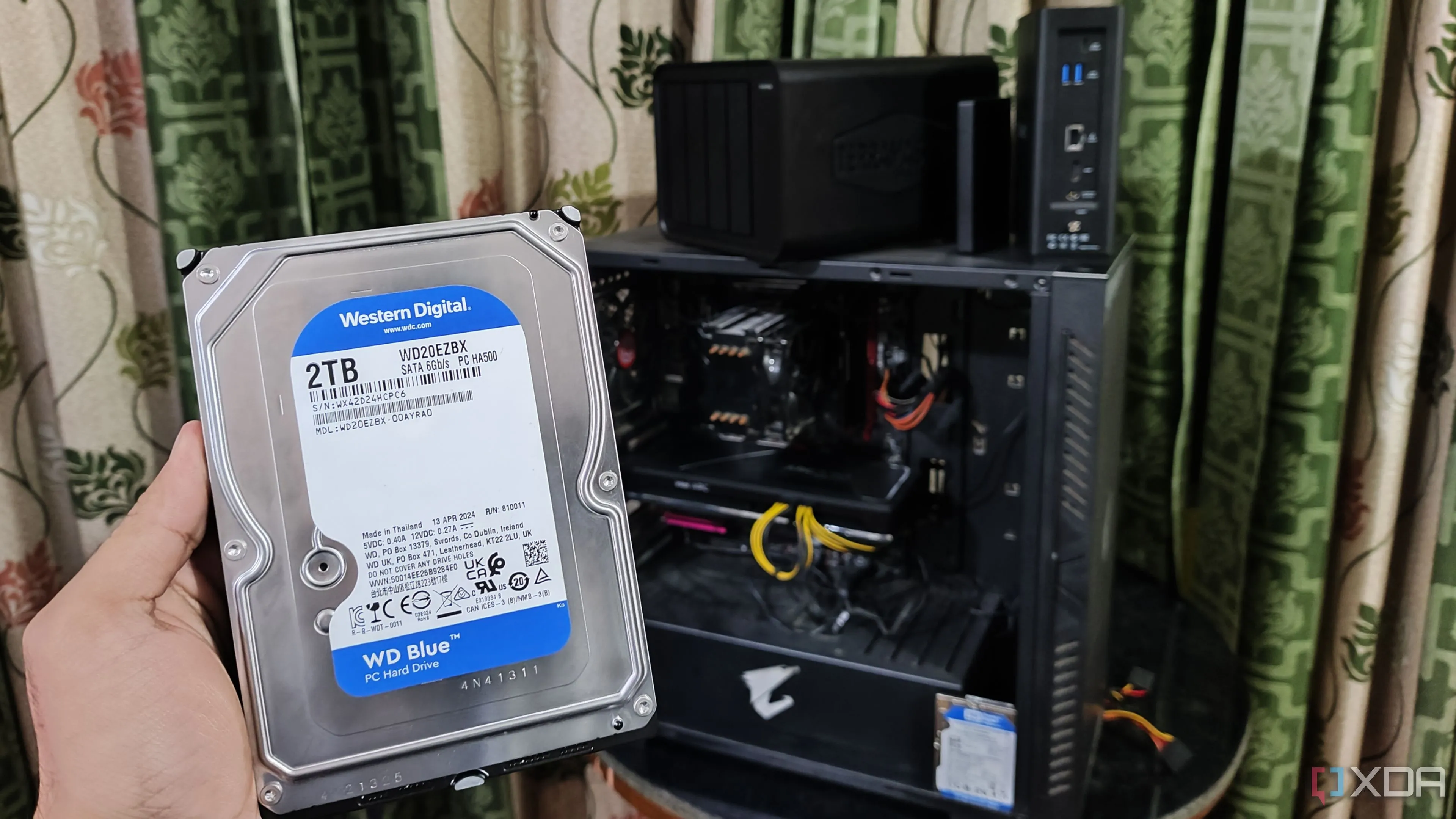 Một người đang cầm ổ cứng trước máy tính và hai thiết bị NAS, minh họa cho hệ thống mạng gia đình và các giải pháp lưu trữ